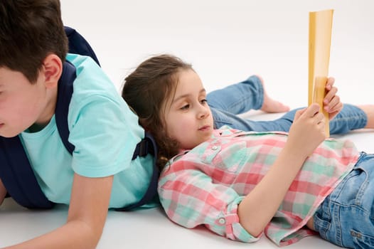 Advertising studio portrait of a beautiful lovely Caucasian little child, preschooler girl lying on the back of her older brother, holding and looking at her workbook isolated on white background