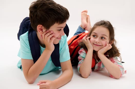 Two Caucasian adorable happy smart school kids, caring older brother and his younger sister with school bags, smiling looking at each other, lying on their belly, isolated on white studio background