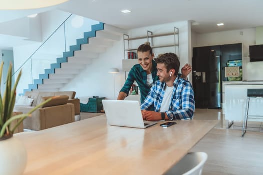 A young married couple is talking to parents, family and friends on a video call via a laptop while sitting in the living room of their modern house.