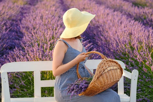 Female donning a straw hat and sundress sits on the bench nestled within a vibrant lavender field, holding a basket with flowers.