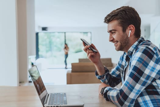 The man sitting at a table in a modern living room, using a smartphone and laptop for business video chat, conversation with friends and entertainment.