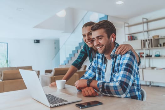 A young married couple is talking to parents, family and friends on a video call via a laptop while sitting in the living room of their modern house.