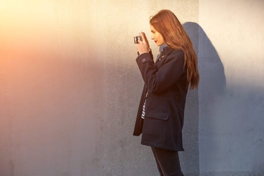 Smiling female photographer in jacket standing in front of wall ready to make new photo. Adorable young brunette woman in trendy outfit posing on concrete wall background with camera