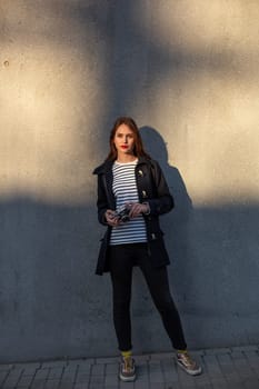 Smiling female photographer in jacket standing in front of wall ready to make new photo. Adorable young brunette woman in trendy outfit posing on concrete wall background with camera