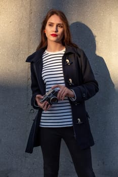 Smiling female photographer in jacket standing in front of wall ready to make new photo. Adorable young brunette woman in trendy outfit posing on concrete wall background with camera