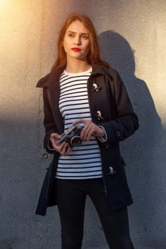 Smiling female photographer in jacket standing in front of wall ready to make new photo. Adorable young brunette woman in trendy outfit posing on concrete wall background with camera