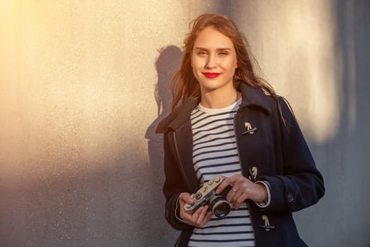 Smiling female photographer in jacket standing in front of wall ready to make new photo. Adorable young brunette woman in trendy outfit posing on concrete wall background with camera