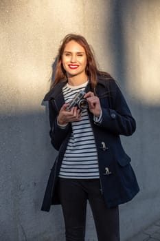 Smiling female photographer in jacket standing in front of wall ready to make new photo. Adorable young brunette woman in trendy outfit posing on concrete wall background with camera