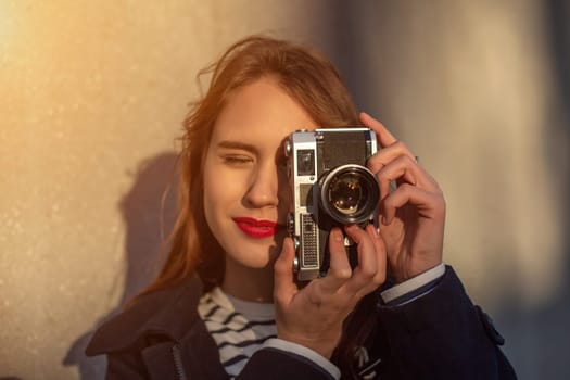 Smiling female photographer in jacket standing in front of wall ready to make new photo. Adorable young brunette woman in trendy outfit posing on concrete wall background with camera