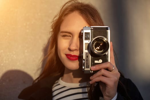 Smiling female photographer in jacket standing in front of wall ready to make new photo. Adorable young brunette woman in trendy outfit posing on concrete wall background with camera