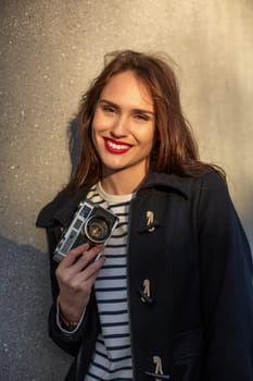 Smiling female photographer in jacket standing in front of wall ready to make new photo. Adorable young brunette woman in trendy outfit posing on concrete wall background with camera