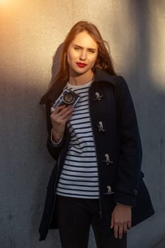 Smiling female photographer in jacket standing in front of wall ready to make new photo. Adorable young brunette woman in trendy outfit posing on concrete wall background with camera