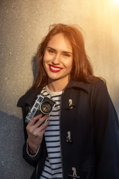 Smiling female photographer in jacket standing in front of wall ready to make new photo. Adorable young brunette woman in trendy outfit posing on concrete wall background with camera