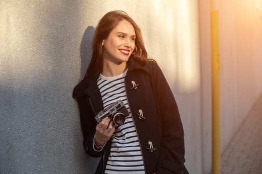 Smiling female photographer in jacket standing in front of wall ready to make new photo. Adorable young brunette woman in trendy outfit posing on concrete wall background with camera