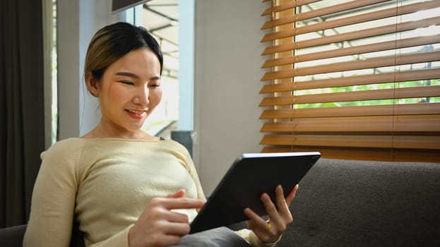 Happy millennial young woman using digital tablet on sofa in living room. People, technology and lifestyle concept.