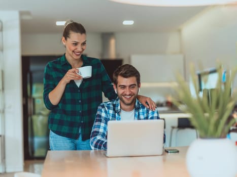 A young married couple is talking to parents, family and friends on a video call via a laptop while sitting in the living room of their modern house.