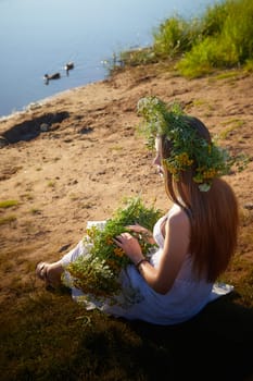 Young brunette girl in a white dress, sundress and wreath of flowers in summer on the coast of river or lake in the evening at sunset. Celebration of the Slavic pagan holiday of Ivan Kupala