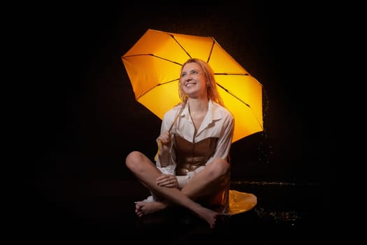 Beautiful sexy skinny girl in a dark hall with splashes of water and yellow umbrella under rain. Female Model blonde woman posing with water on black background in the studio