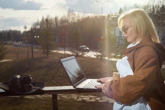 Fashionable beautiful blonde girl is working with laptop in gazebo on sunny autumn, spring day. Businesswoman, entrepreneur, student, manager works outdoors against the backdrop of natural landscape