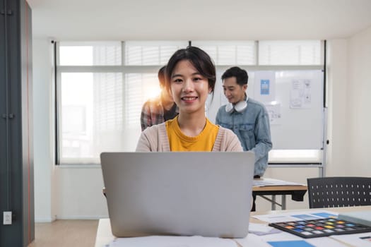 young Asian female marketing assistant is working on her work on her laptop at her desk while her coworkers are working in the room