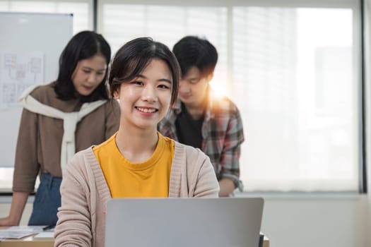 young Asian female marketing assistant is working on her work on her laptop at her desk while her coworkers are working in the room