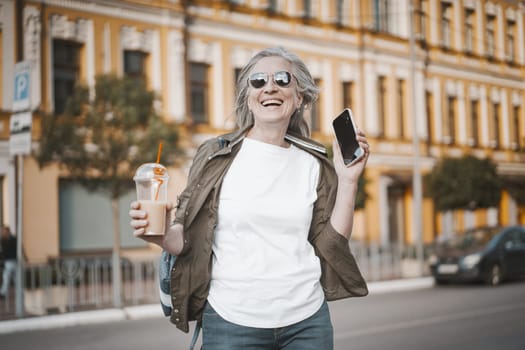 Happy senior old woman smiling outdoors. She speaking on phone, engaging in modern communication, while enjoying refreshing drink from transparent plastic cup. Mature lady in moment of joy. High quality photo