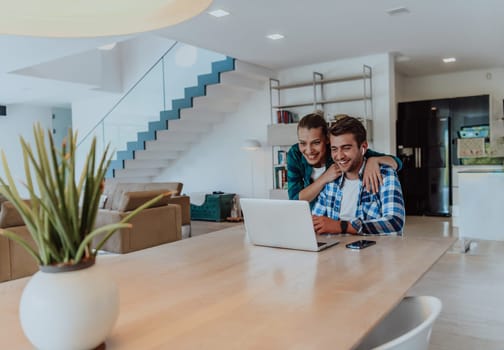 A young married couple is talking to parents, family and friends on a video call via a laptop while sitting in the living room of their modern house.