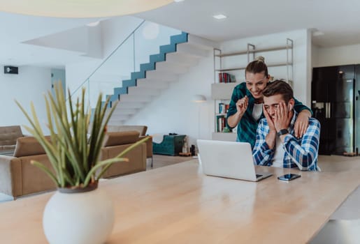 A young married couple is talking to parents, family and friends on a video call via a laptop while sitting in the living room of their modern house.