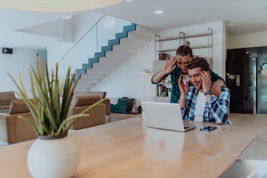 A young married couple is talking to parents, family and friends on a video call via a laptop while sitting in the living room of their modern house.