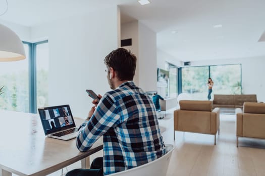 The man sitting at a table in a modern living room, using a smartphone and laptop for business video chat, conversation with friends and entertainment.