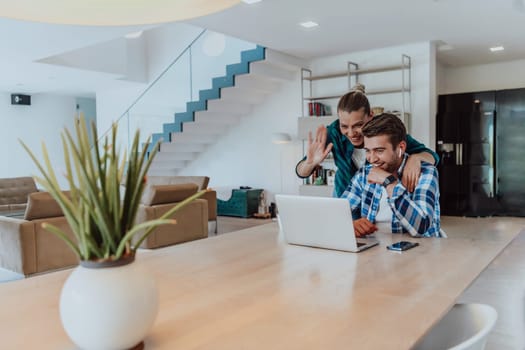 A young married couple is talking to parents, family and friends on a video call via a laptop while sitting in the living room of their modern house.