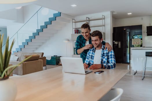 A young married couple is talking to parents, family and friends on a video call via a laptop while sitting in the living room of their modern house.