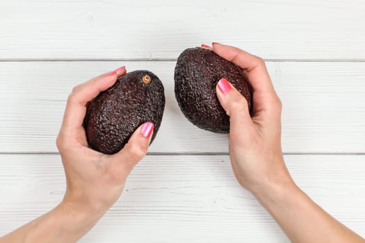 Top down view, woman hands with pink nails holding two whole dark ripe avocados over white boards desk.