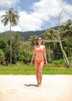 Young woman in red bikini and sunglasses walking on the beach, jungle with palm trees behind her.