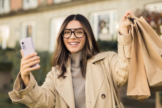 Fashionable beautiful model woman 30s in trendy glasses holding up paper shopping bags and phone happily smiling.