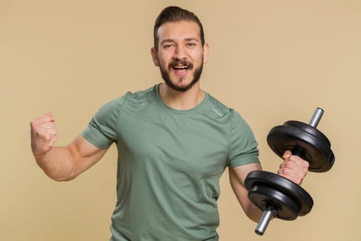 Excited athlete sportive man shouting, celebrating success, winning, goal achievement, good news, lottery luck, wow reaction. Middle eastern sportsman guy weightlifting dumbbell on beige background
