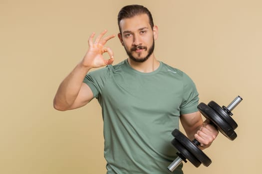 Okay. Athlete sportive man looking at camera showing ok gesture, like sign positive, approve something good, feedback, celebrate, agree. Middle eastern sportsman guy with dumbbell on beige background