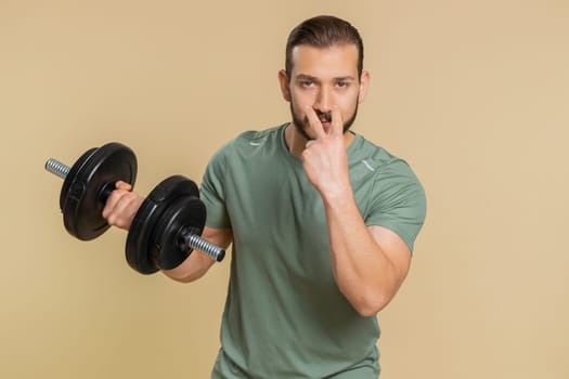 I am watching you. Athlete sportive man pointing at eyes and camera, show I am watching you gesture, spying on someone start working out. Middle eastern sportsman guy with dumbbell on beige background