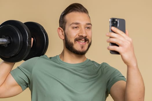 Athlete sportive man texting share messages content on smartphone social media applications online, sms message, browsing, reading news. Middle eastern sportsman guy with dumbbell on beige background