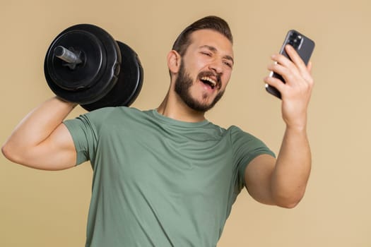 Athlete sportive man use mobile smartphone typing browsing say Wow yes found out great big win good news celebrate doing winner gesture. Middle eastern sportsman guy with dumbbell on beige background