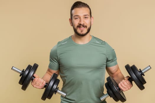 Athlete young sportive man working out. Middle eastern bodybuilder instructor sportsman guy training biceps, weightlifting dumbbells indoors on studio beige background. Strong, exercise, motivation