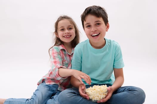Caucasian happy cute children, preteen boy and little girl, brother and sister sitting over white isolated studio background, watching cartoons or movie, eating popcorn. kids. Lifestyle. Entertainment