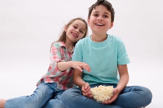 Caucasian amazed funny children, school kids, handsome teen boy and lovely little girl, brother and sister, having fun together, smiling, watching cartoons and eating popcorn. Lifestyle. Entertainment