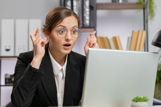 Amazed business woman girl in glasses crossed fingers looking at laptop screen asking for good luck news wishing good exam results dreaming about win victory in lottery jackpot, money profit at office