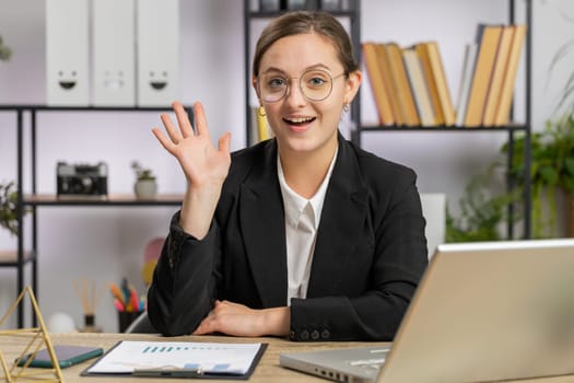 Hello. Caucasian business woman working on laptop computer smiling friendly at camera, waving hands gesturing hi greeting or goodbye, welcoming with hospitable expression at home office workplace desk