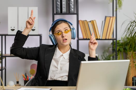 Happy relaxed overjoyed Caucasian business woman working on laptop computer at office wearing headphones listening favorite energetic disco music and dancing. Freelancer girl relaxing, taking a break