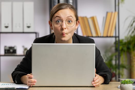 Caucasian joyful business woman hiding behind laptop computer making funny face, fooling around, remote working disrespecting someone at office workplace. Professional freelancer girl