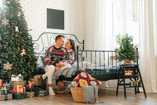 Young woman and man sitting on bed in sweaters and hugging near Christmas tree close up