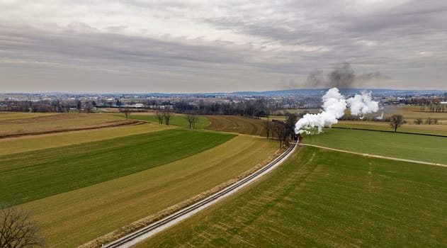 An Aerial View of a Steam Passenger Train Approaching, Traveling Thru Open Farmlands, Blowing Lots of White Smoke, on a Winter Day