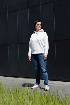 Full Length Sportive Mature Brunette Woman in White Hoody, Blue Jeans and Sunglasses Looks Away, Dark Granite Wall On Background. Confident 40 Yo Beautiful Caucasian Woman in City. Vertical Plane.